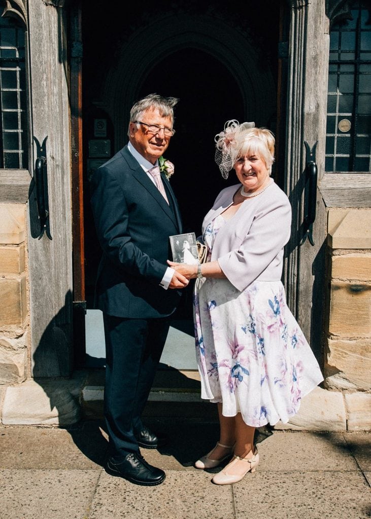 Groom parents holding their wedding photo in the same spot outside of St Cuthberts Church in Blyth